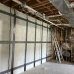 Construction site interior featuring exposed wooden beams, metal ducts, a ladder, and a textured white wall with invisibeam foundation support straps installed and anchored to the concrete slab floor.