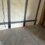 Interior view of a basement wall featuring invisibeam foundation support straps anchored to the floor and black waterproofing paint on cinder blocks, with a concrete floor and unfinished wood framing.