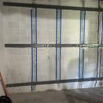 A partially treated basement wall with invisibeam foundation support straps and blue painter's tape, showing signs of moisture and a ladder nearby.