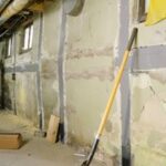 A man inspects a basement with exposed walls, a shovel leaning against the wall, and a ladder in a dimly lit corner.