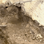 Close-up of muddy excavation revealing a gray pipe, dirt, and a small water pool near a textured wall, indicating plumbing work.