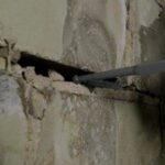 A close-up of a wall showing a crack, with a metal tool inserted into the gap for repair or inspection purposes.