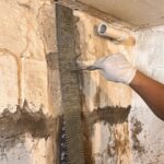 A gloved hand applies material to a reinforced wall with Invisibeam in a basement setting, showcasing home repair work.