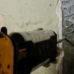 A caulking gun applying adhesive to a mesh tape on a textured wall, with peeling paint and exposed surface visible.