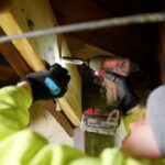 A person in a gray beanie and neon yellow jacket uses a drill to secure a wooden beam in a dimly lit space.