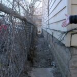 Narrow alleyway beside a building, with a person gesturing; fence with tangled wires and overgrown weeds visible, cluttered ground.