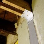 Close-up of a metal bracket securing wooden beams to a concrete wall in a basement setting, showcasing structural support.