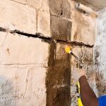 A worker uses a torch to repair a cracked concrete wall in a basement, showcasing damage and restoration efforts.