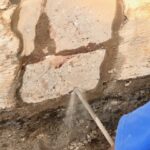 A worker in a blue shirt uses a spray tool to apply a repair mixture to a damaged section of a wall with peeling paint and cracks.