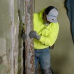 A worker in a neon yellow jacket and cap supports a rusty metal beam in a partially renovated room next to a ladder.