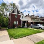 A charming brick home with a steeply pitched roof, lush front lawn, and parked car, set among trees and neighboring houses.