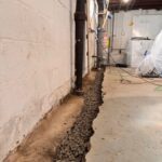 A narrow trench filled with gravel runs alongside a black pipe in a basement, with visible construction tools and plastic sheeting nearby.