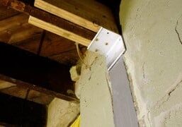 Close-up of a metal bracket securing wooden beams to a concrete wall in a basement setting, showcasing structural support.