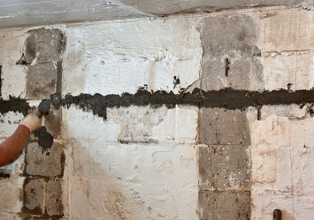 A worker applies cement to a partially finished wall, showcasing uneven surfaces and grey concrete blocks in a dimly lit basement.