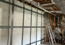 Construction site interior featuring exposed wooden beams, metal ducts, a ladder, and a textured white wall with invisibeam foundation support straps installed and anchored to the concrete slab floor.